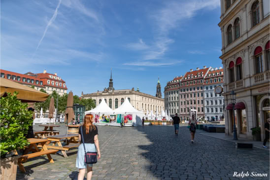 Eine gepflasterte Straße mit Menschen, die spazieren gehen, umgeben von historischen Gebäuden und einem weißen Zelt im Hintergrund unter einem blauen Himmel.